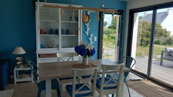 A dining area with a wooden table and white chairs, a white cabinet with glass doors, and a view of the outdoors. - Maison Bord de mer (Treffiagat)