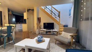 Een woonkamer met een televisie, een eethoek met stoelen, een trap en een keuken op de achtergrond.