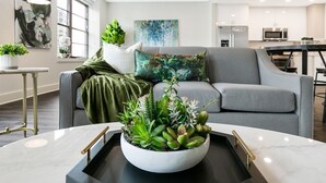 Una sala de estar moderna con un sofá gris, una mesa de centro blanca con una bandeja negra, y una variedad de plantas verdes.