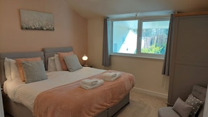 A bedroom with a large bed, a nightstand, a chair, and a window with a view of buildings. - Holly -  a dunley cottages - devon that sleeps 2 guests  in 1 bedroom (Bovey Tracey)