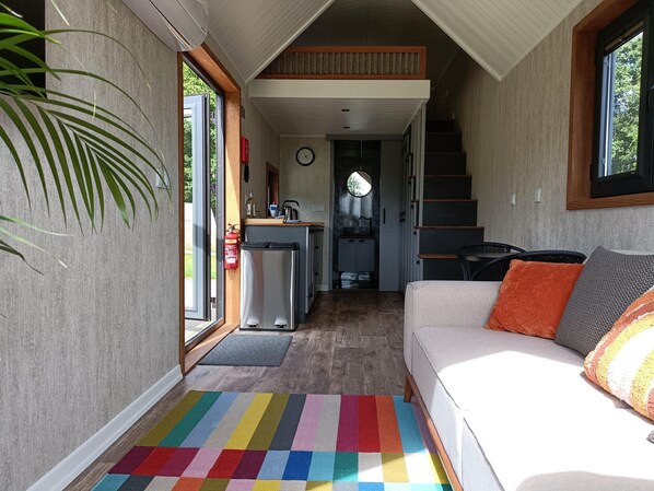 Un espacio habitable compacto con sofá, una alfombra colorida, una zona de cocina y una escalera que sube a un nivel superior.