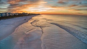 Una playa con cielo despejado y mar tranquilo, edificios al fondo y un camino visible a lo largo de la orilla.