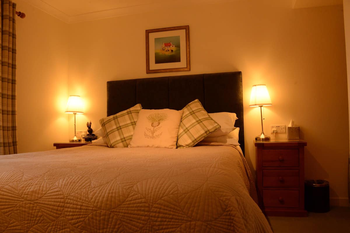 A neatly made bed with a quilt, two bedside lamps, a framed picture on the wall, and a small nightstand with a lamp and a trash can.