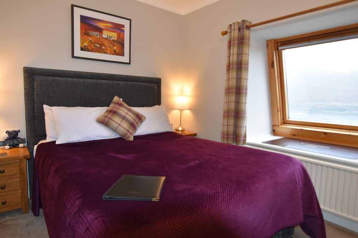 A neatly made bed with a purple blanket, a bedside lamp, a framed picture on the wall, and a window with a view.