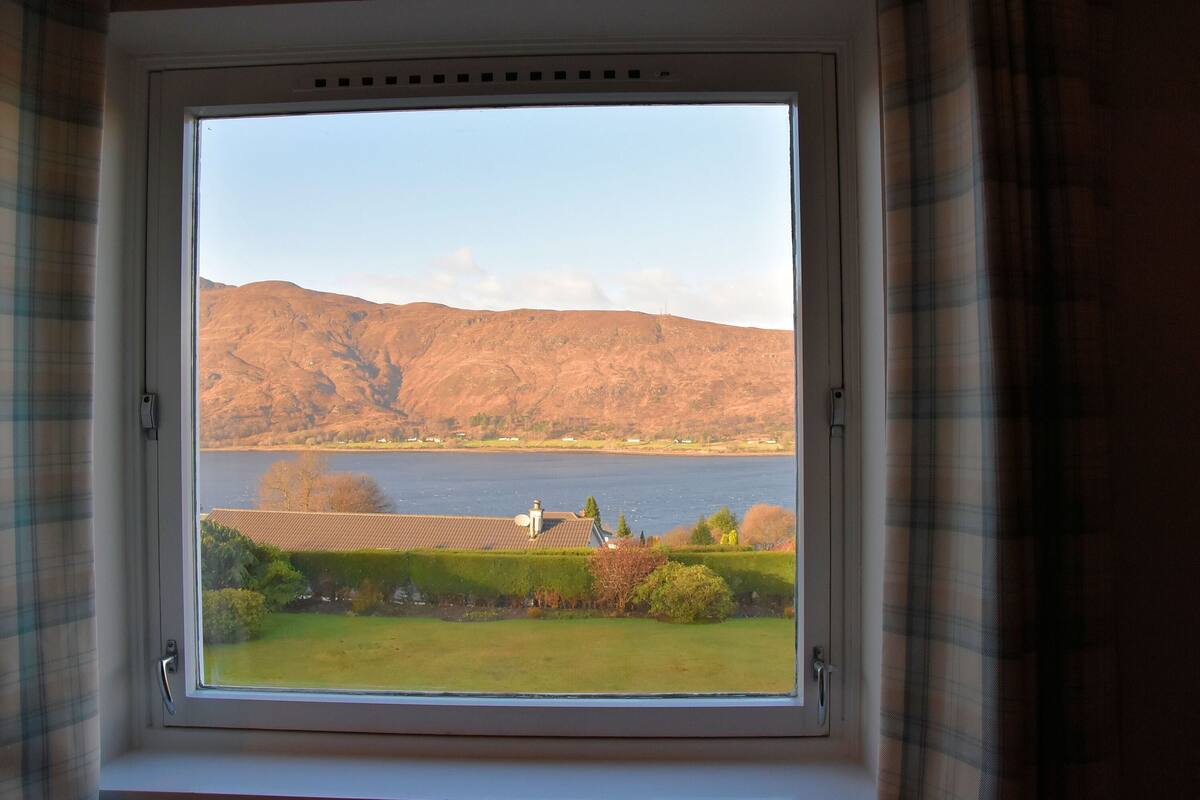 A window with a view of a lake, hills, and a building with a tiled roof.