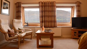A living room with two chairs, a table, a television, and a window with curtains. - Lochview Guest House (Fort William)