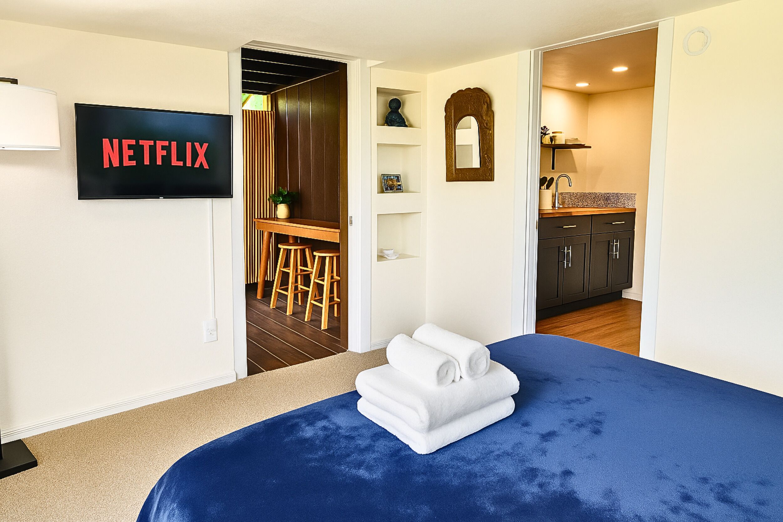 A hotel room with a bed, a TV displaying 'NETFLIX', a small kitchen area, and a bathroom.