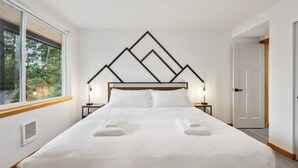 Ein modernes Schlafzimmer mit einem großen Bett, einem geometrischen Kopfteil, zwei Nachttischen mit Lampen und Blick auf Grün durch ein Fenster.