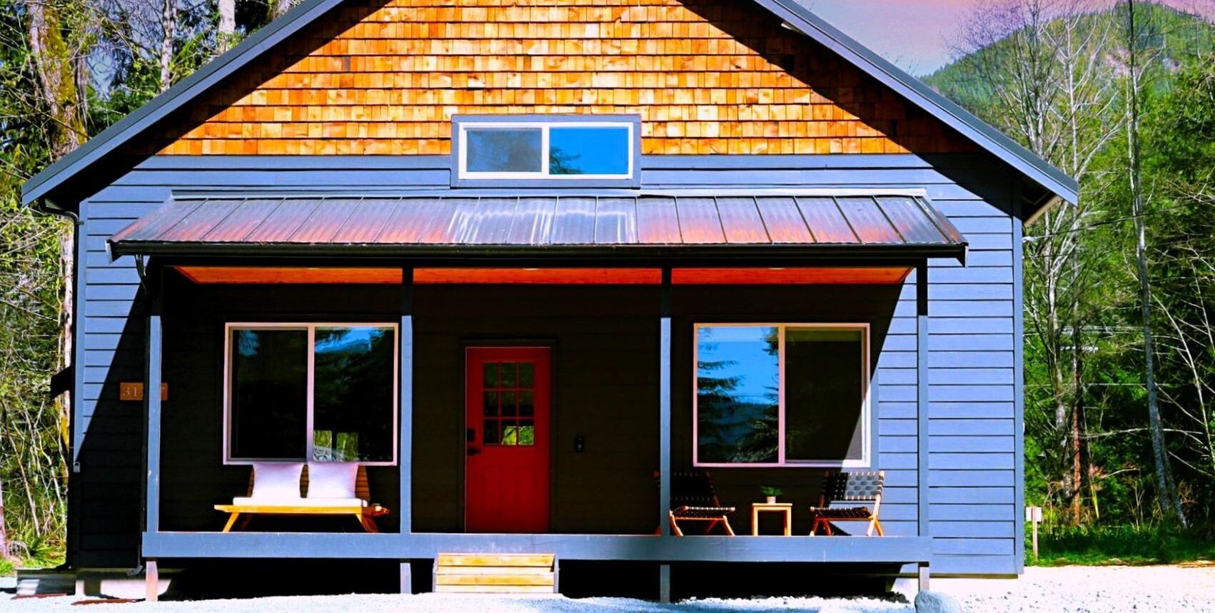 A cabin with a red door, two large windows, and a porch with outdoor seating.