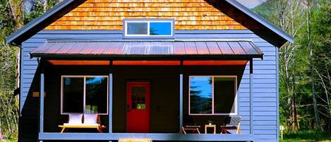 A cabin with a red door, two large windows, and a porch with outdoor seating.