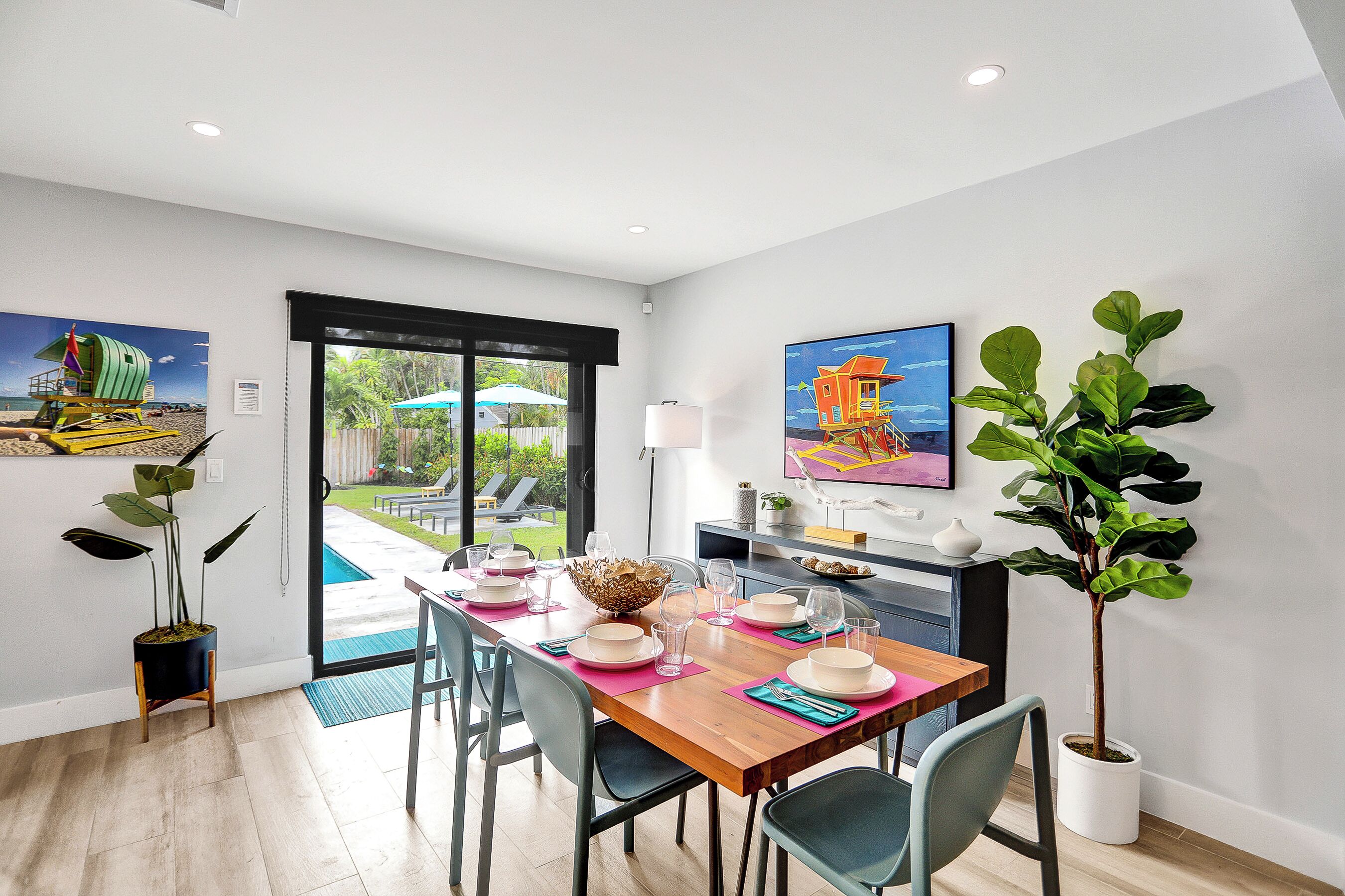 A dining area with a wooden table set for four, surrounded by chairs, with a view of a pool and garden through a glass door.