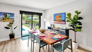 A dining area with a wooden table set for four, surrounded by chairs, with a view of a pool and garden through a glass door.