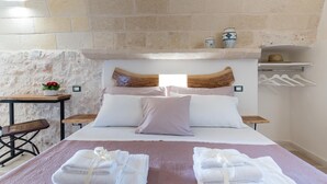 A bedroom with a stone wall, a wooden headboard, a bed with white and pink bedding, a small table with a vase and flowers, and a chair. - 3419 Trullo Mest Marcucc by Perle di Puglia (San Michele Salentino)