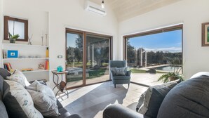 A modern living room with a sofa, a chair, a bookshelf, and a large sliding glass door leading to an outdoor patio. - 3419 Trullo Mest Marcucc by Perle di Puglia (San Michele Salentino)
