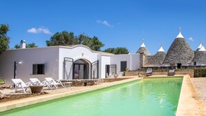 A pool area with lounge chairs, a stone building with conical roofs, and a white building with an arched doorway. - 3598 Trullo Yoram by Perle di Puglia (Francavilla Fontana)