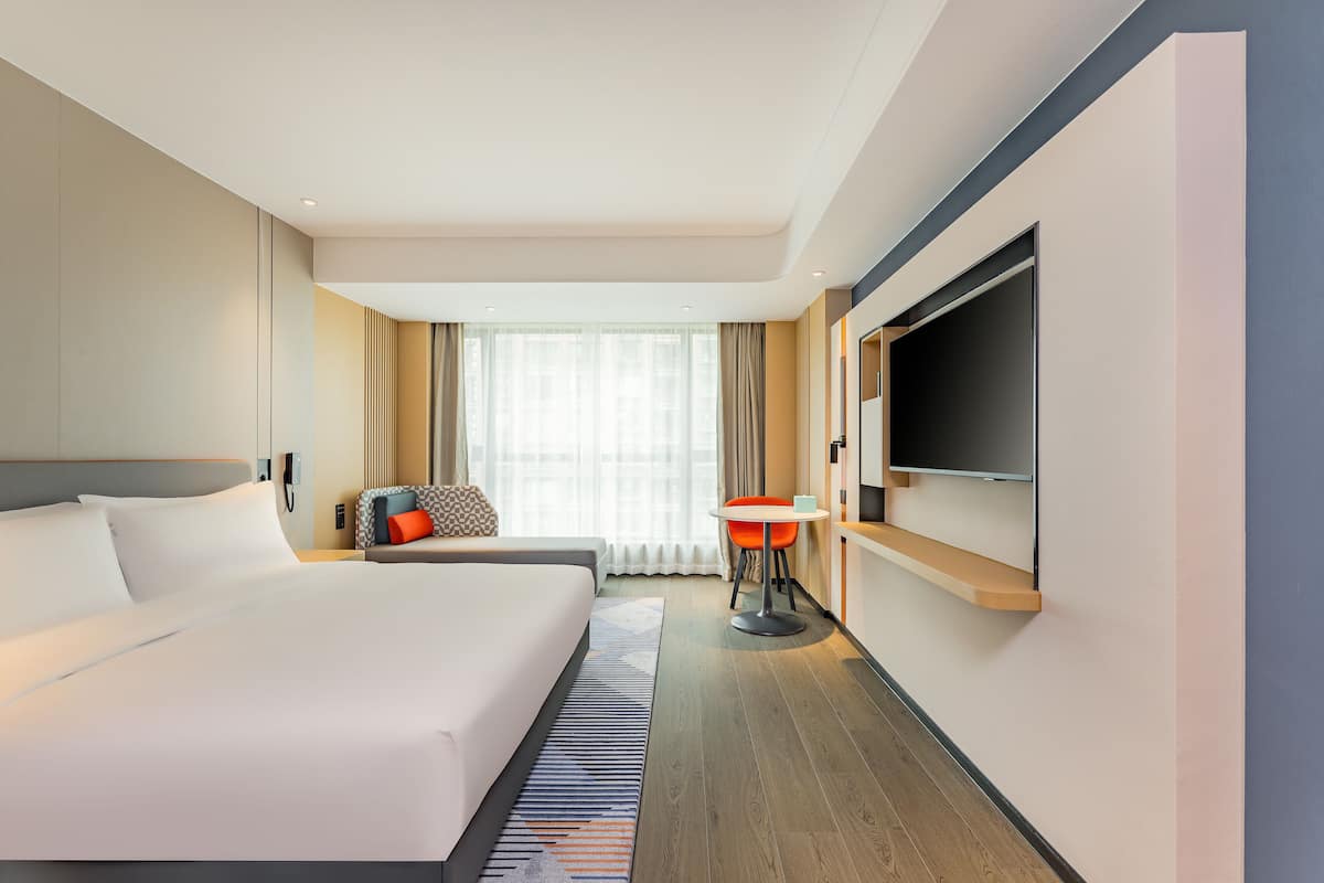 Habitación de hotel con una cama grande, televisión montada en la pared, una mesita y una ventana con cortinas.