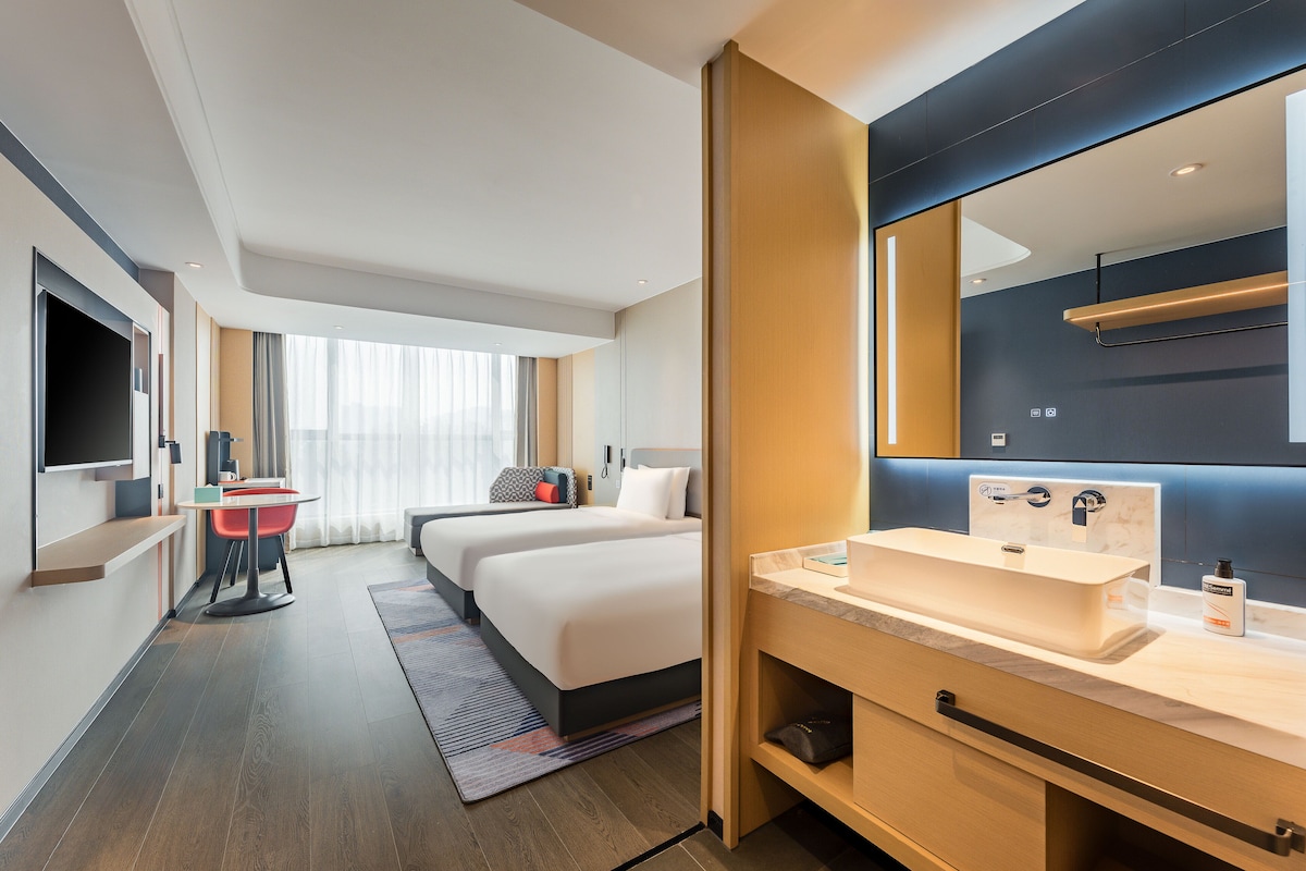 A modern hotel room with a large bed, a desk with a chair, a large mirror, and a bathroom with a sink and mirror.