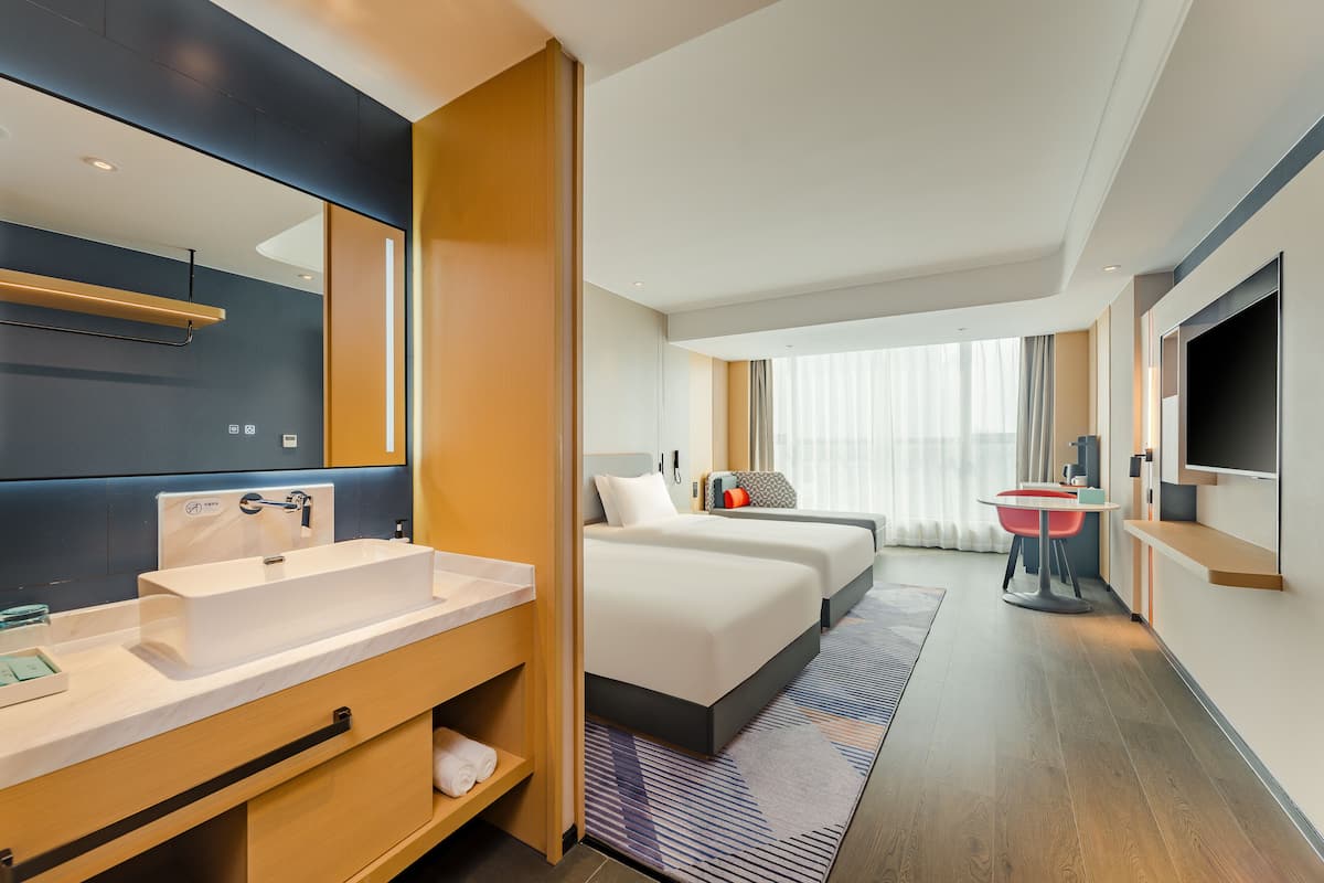 Una habitación de hotel moderna con un espejo grande, un lavamanos blanco, una mesita de noche, un televisor de pantalla plana y un amplio ventanal con cortinas.
