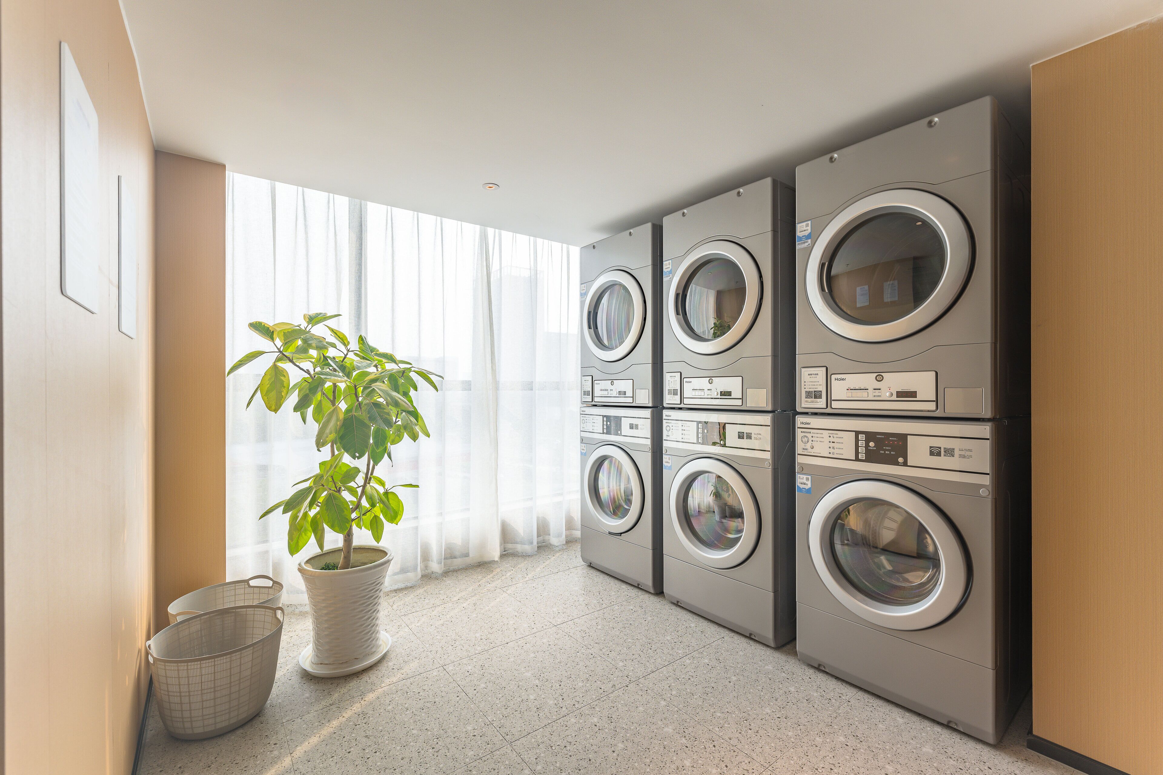 A laundry room with four front-loading washers and dryers, a potted plant, and a trash bin.