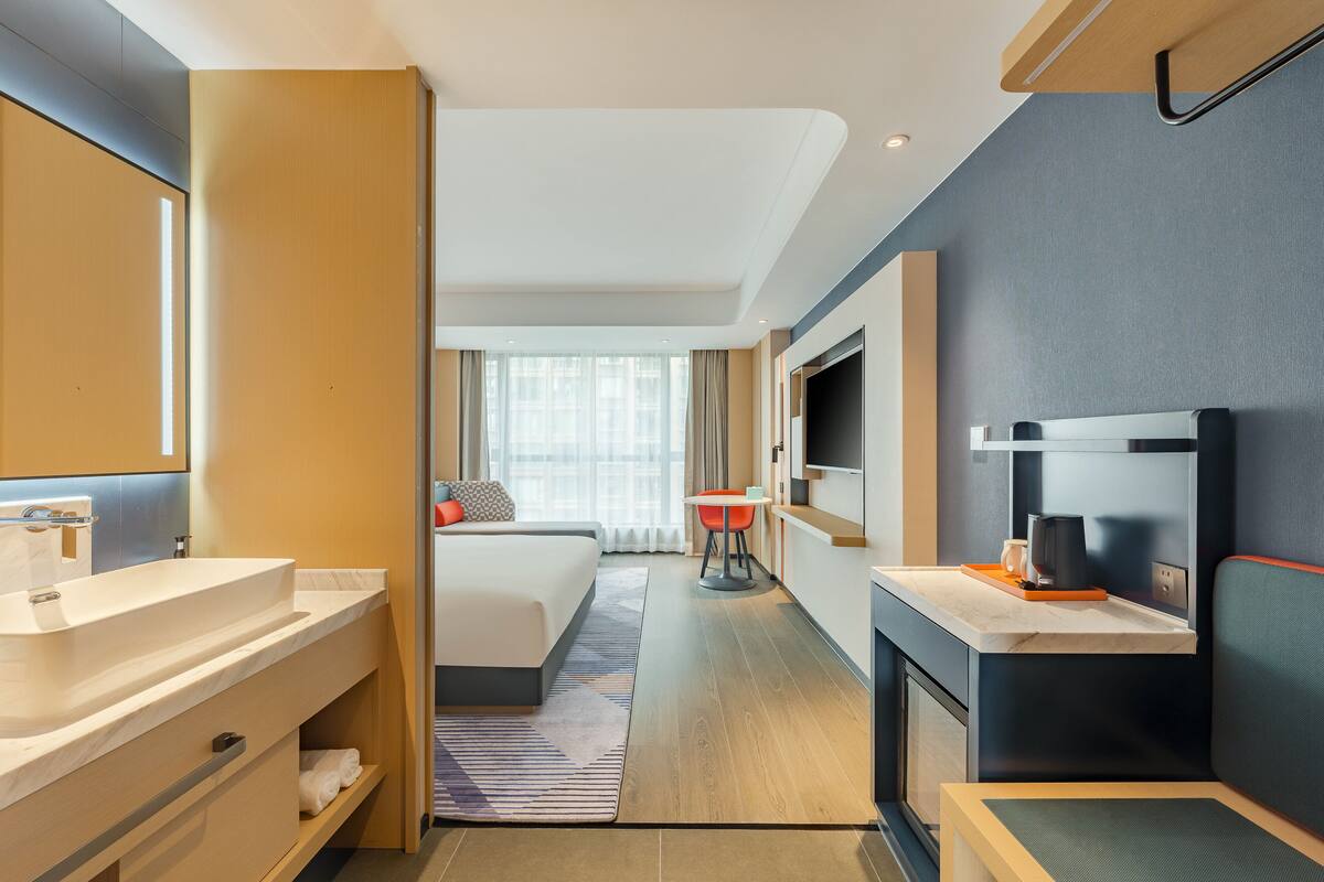 Una habitación de hotel moderna con una cama grande, un baño con lavamanos y espejo, una pequeña mesa de comedor y un televisor.