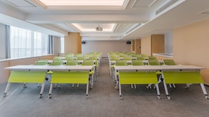 Un salón de conferencias con filas de sillas verdes, una mesa blanca y un proyector en el techo.