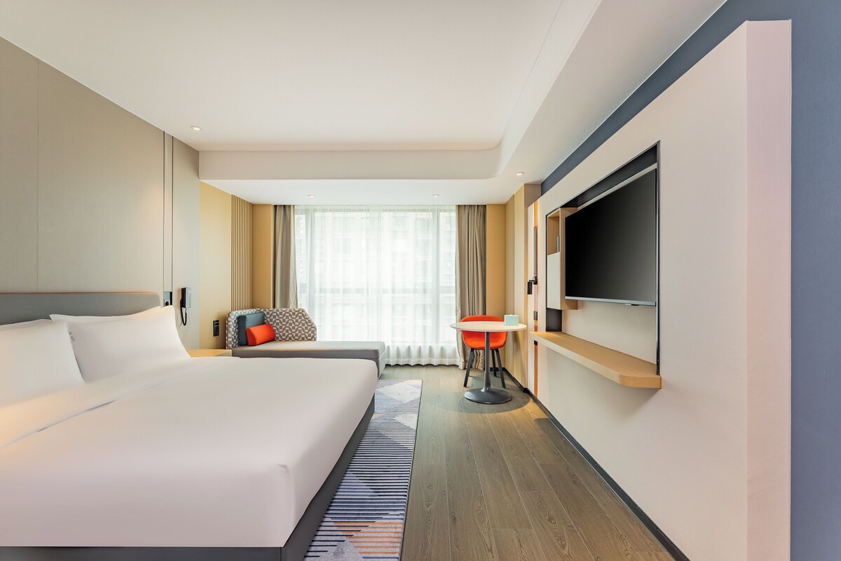 Hotelkamer met een groot bed, een aan de muur bevestigde televisie, een kleine tafel en een raam met gordijnen.