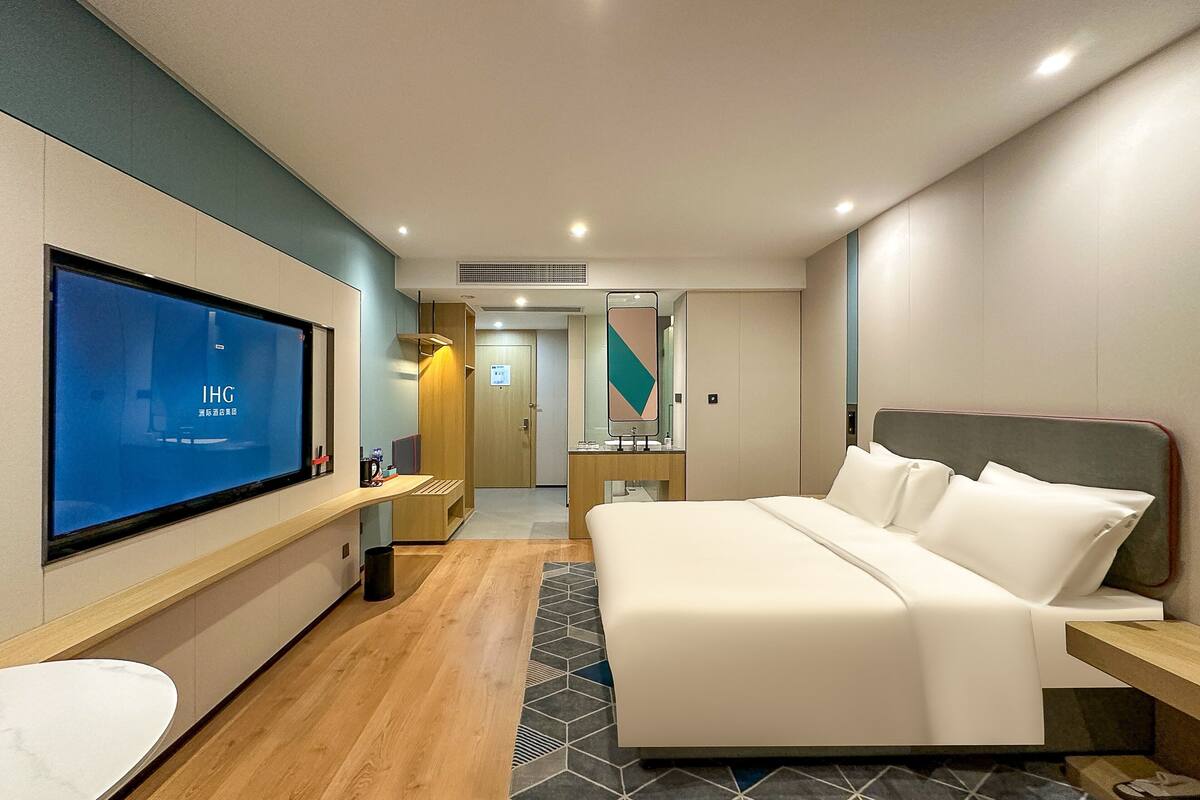 A modern hotel room with a large bed, a TV on the wall, a desk, and a bathroom.