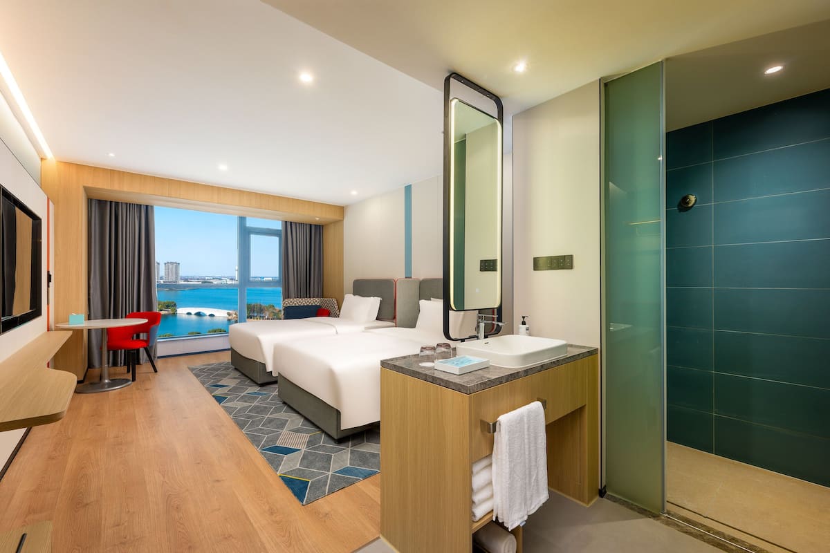 A hotel room with a bed, a desk, a bathroom with a mirror, and a view of the city.