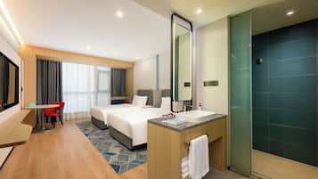 A hotel room with a bed, a desk, a chair, a bathroom with a sink and mirror, and a shower area.