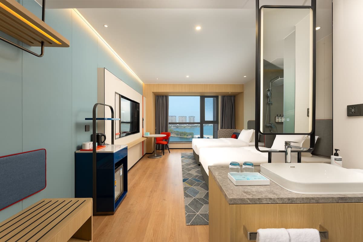A modern hotel room with a large bed, a desk with a coffee machine, a seating area with a red chair, a large window with a city view, and a separate bathroom with a large mirror and sink.