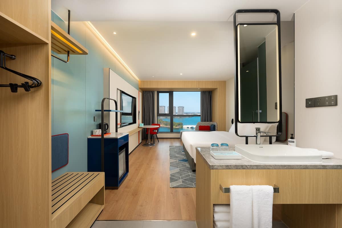 A modern hotel room with a large bed, a bathroom with a sink and mirror, a small seating area with a table and chairs, and a view of the city through large windows.