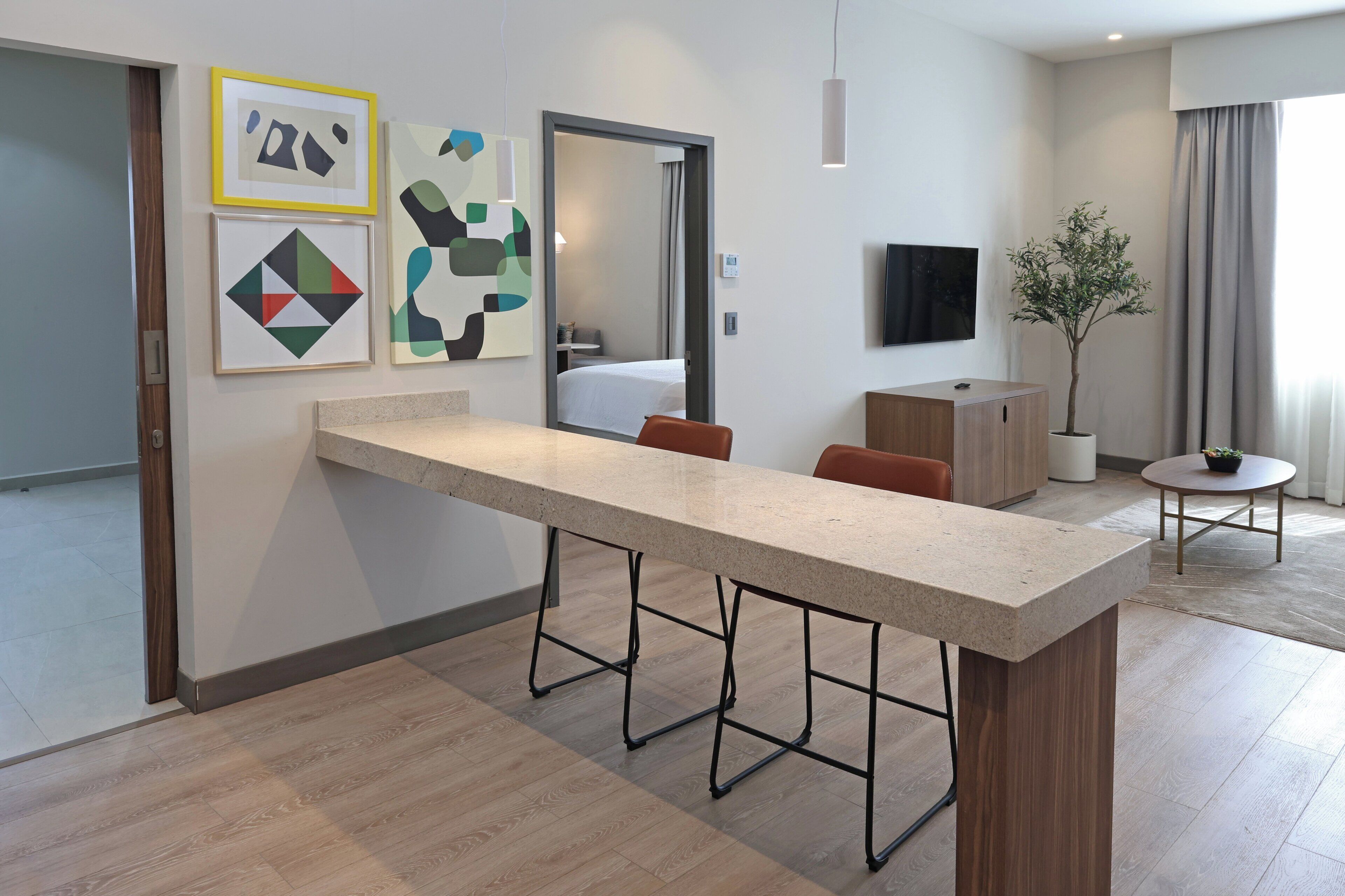 A modern hotel room with a long counter, a bed in the background, a TV, and a potted plant.