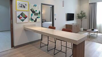 A modern hotel room with a long counter, a bed in the background, a TV, and a potted plant.