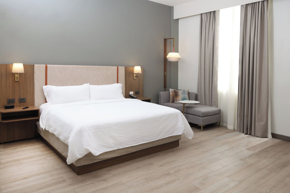 A modern hotel room with a large bed, bedside tables, a small sofa, and a large window with curtains.