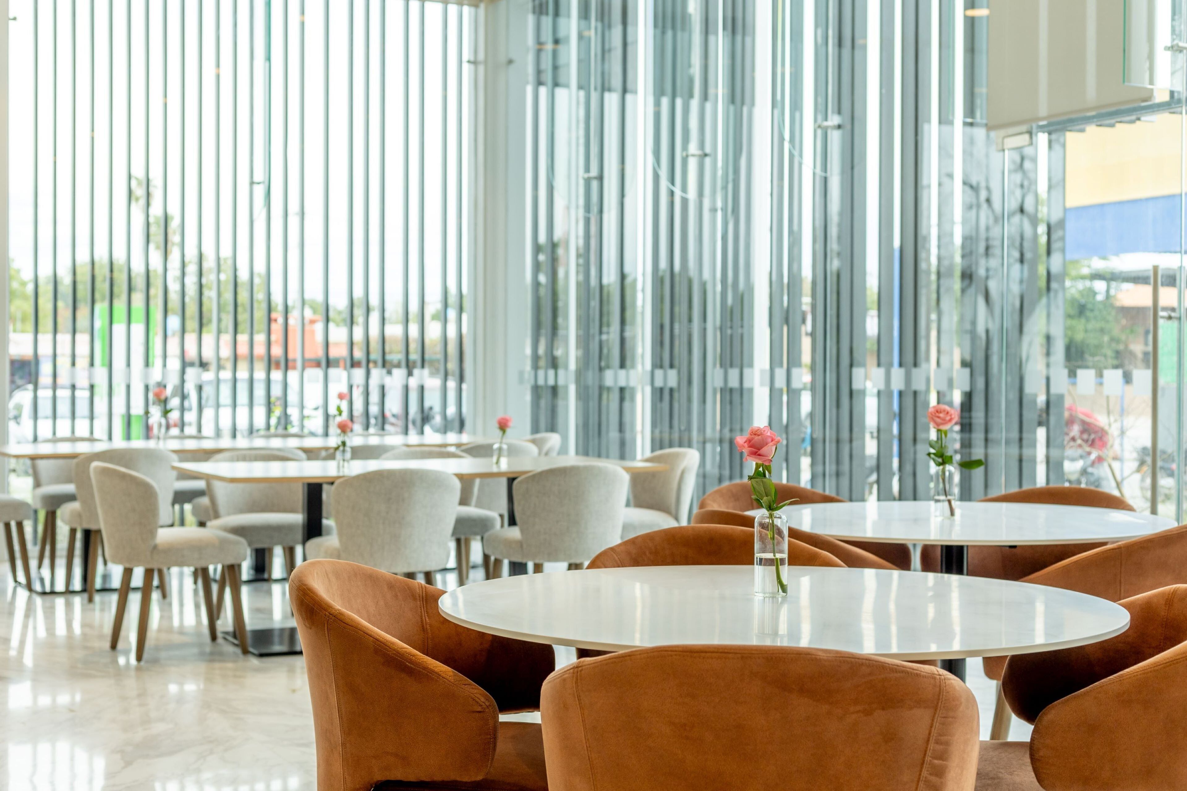 A modern dining area with round tables and cushioned chairs, large windows with vertical blinds, and a view of the outside.