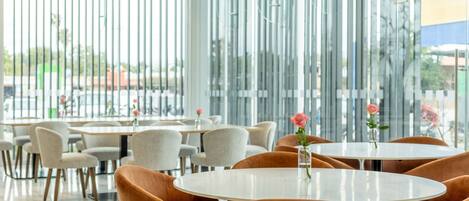 A modern dining area with round tables and cushioned chairs, large windows with vertical blinds, and a view of the outside.