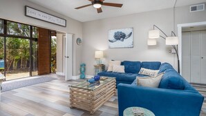 A modern living room with a blue sofa, a glass coffee table, and a ceiling fan. - Harbor Towers 1st Fl. West Facing (Siesta Key)