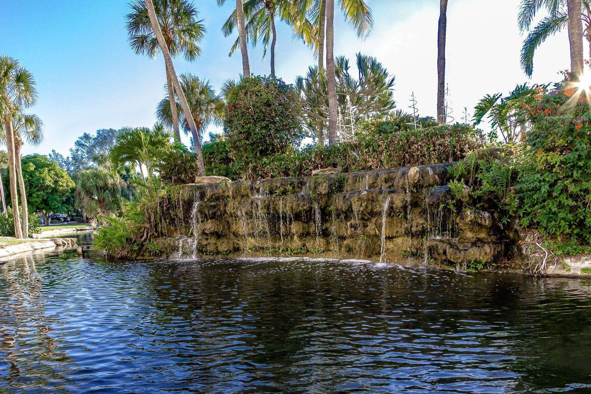 A waterfall cascading over a rocky ledge into a calm pond, surrounded by lush greenery and palm trees.