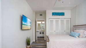 A bedroom with a bed, a TV, a closet, and a hallway leading to a bathroom. - Harbor Towers 1st Fl. West Facing (Siesta Key)