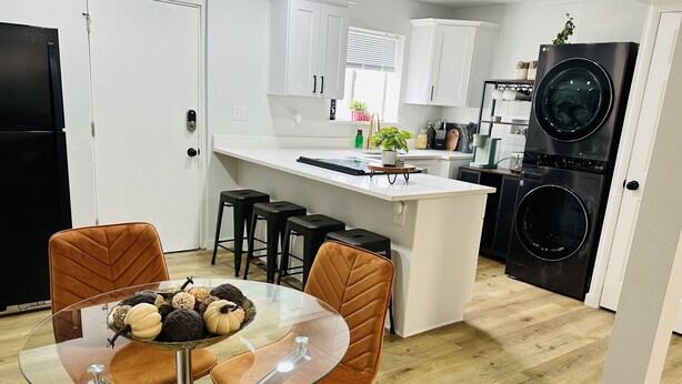 A modern kitchen with white cabinets, a black refrigerator, a built-in washing machine, and a glass dining table with orange chairs. - Home sweet home in West Jordan (West Jordan)