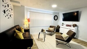 A modern living room with a black leather sofa, a brown armchair, and a white coffee table. A flat-screen TV is mounted on the wall, and a window with blinds is visible. - Home sweet home in West Jordan (West Jordan)