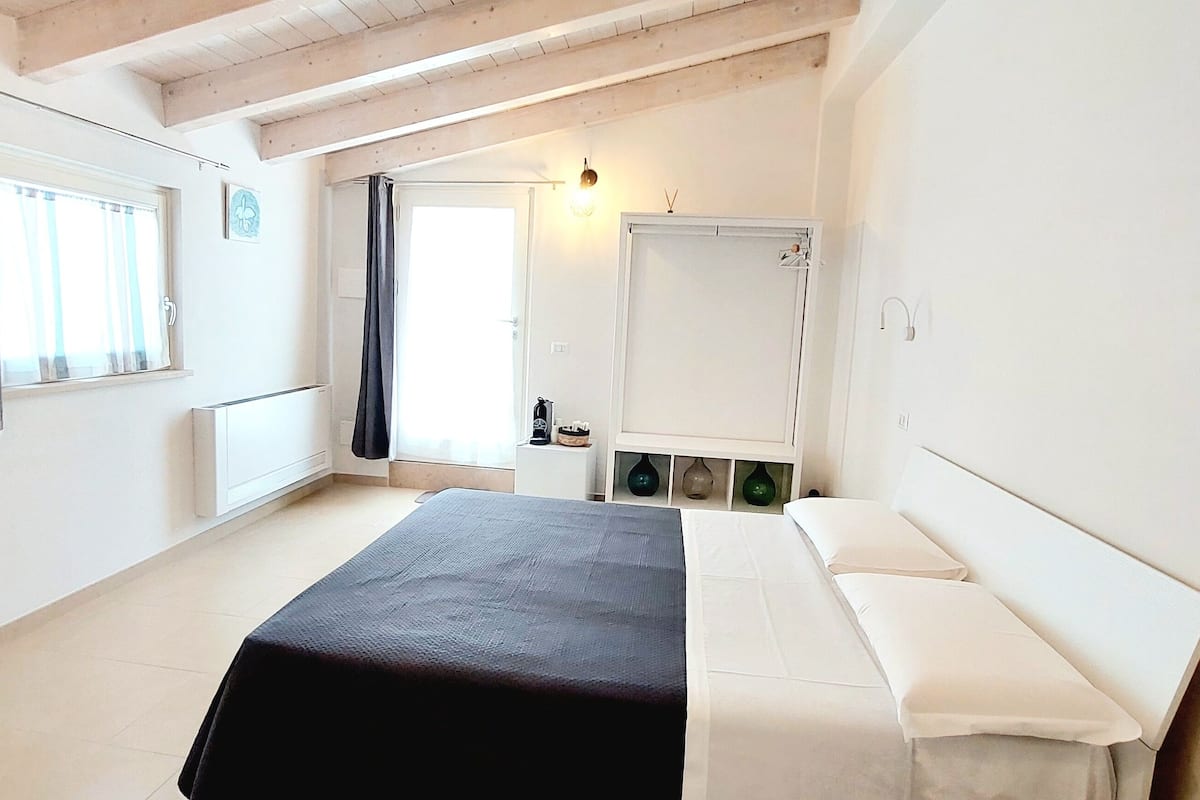 Ett sovrum med en säng, en spegel, ett fönster med gardiner, ett sängbord och en vägglampa.