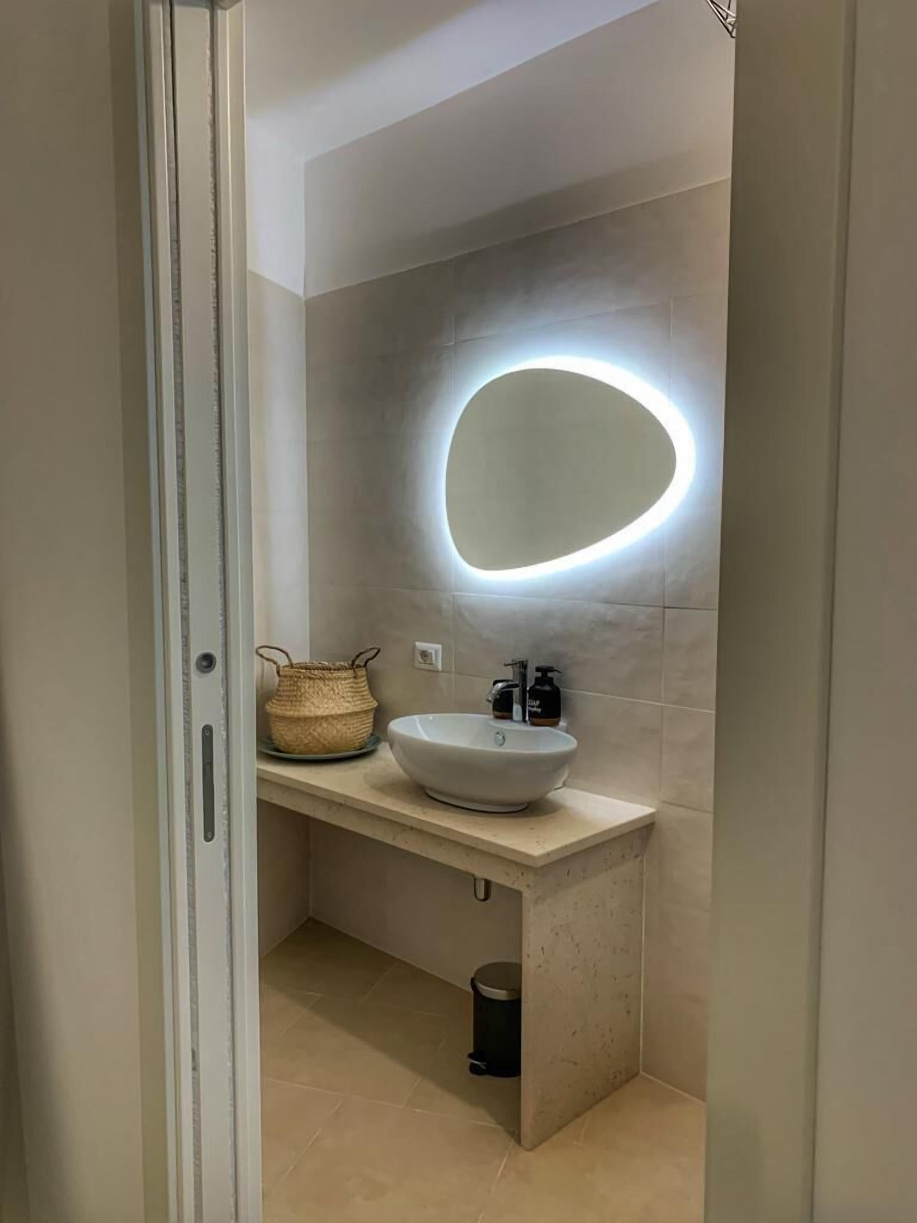 A bathroom with a white sink, a round mirror with a light, a woven basket, and a trash bin.