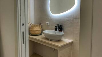 A bathroom with a white sink, a round mirror with a light, a woven basket, and a trash bin.