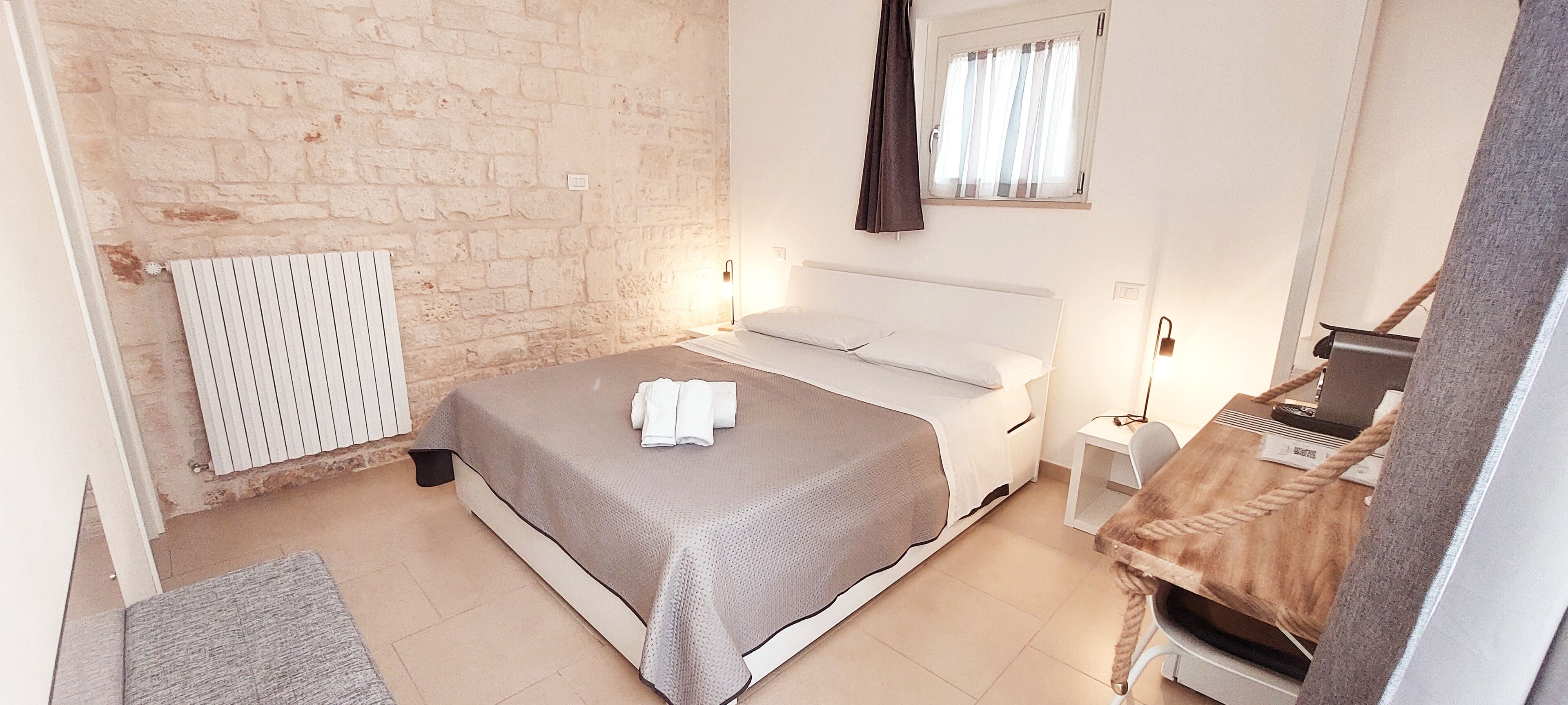 A bedroom with a bed, a radiator, a window with curtains, a bedside table, and a small stool.