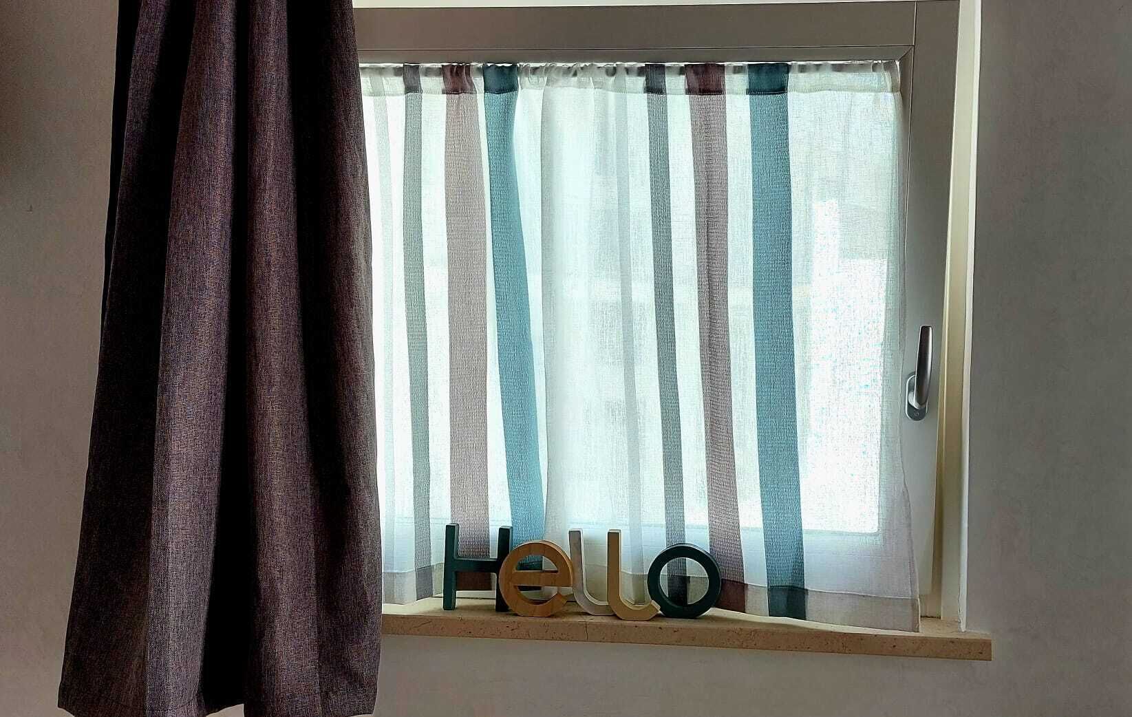 Een raam met gestreepte gordijnen en een houten plankje waarop de tekst 'Hello' staat.