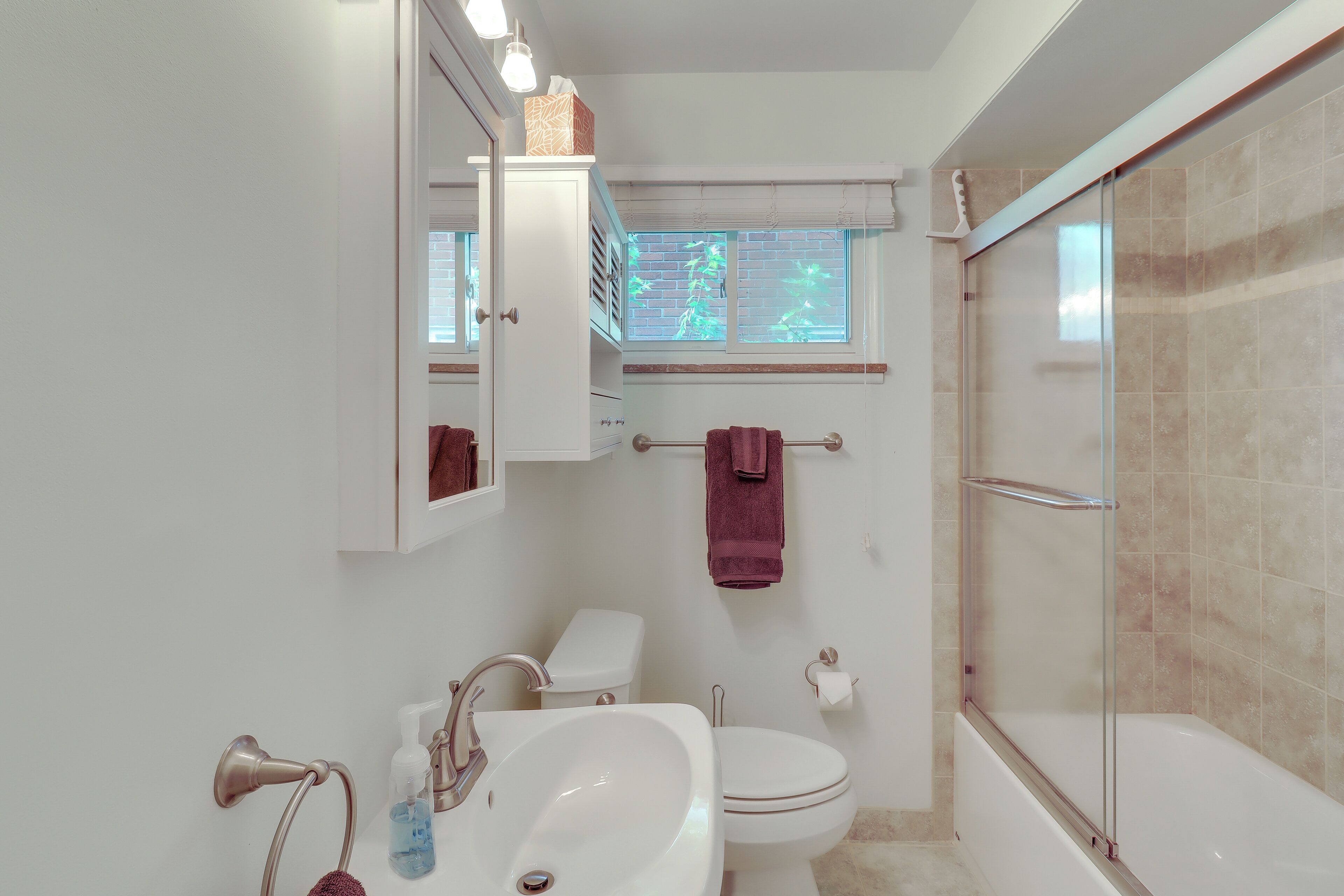 A bathroom with a white sink, toilet, and bathtub. There is a window with blinds, a towel rack, and a mirror above the sink.