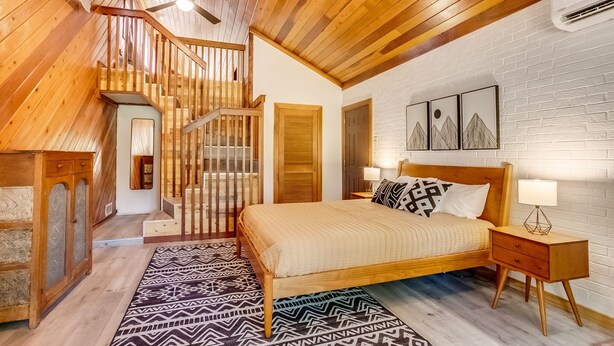 Ein Schlafzimmer mit einem hölzernen Bett, Nachttischen, einem gemusterten Teppich, einer Treppe und einer weißen Ziegelwand mit gerahmten Bildern.