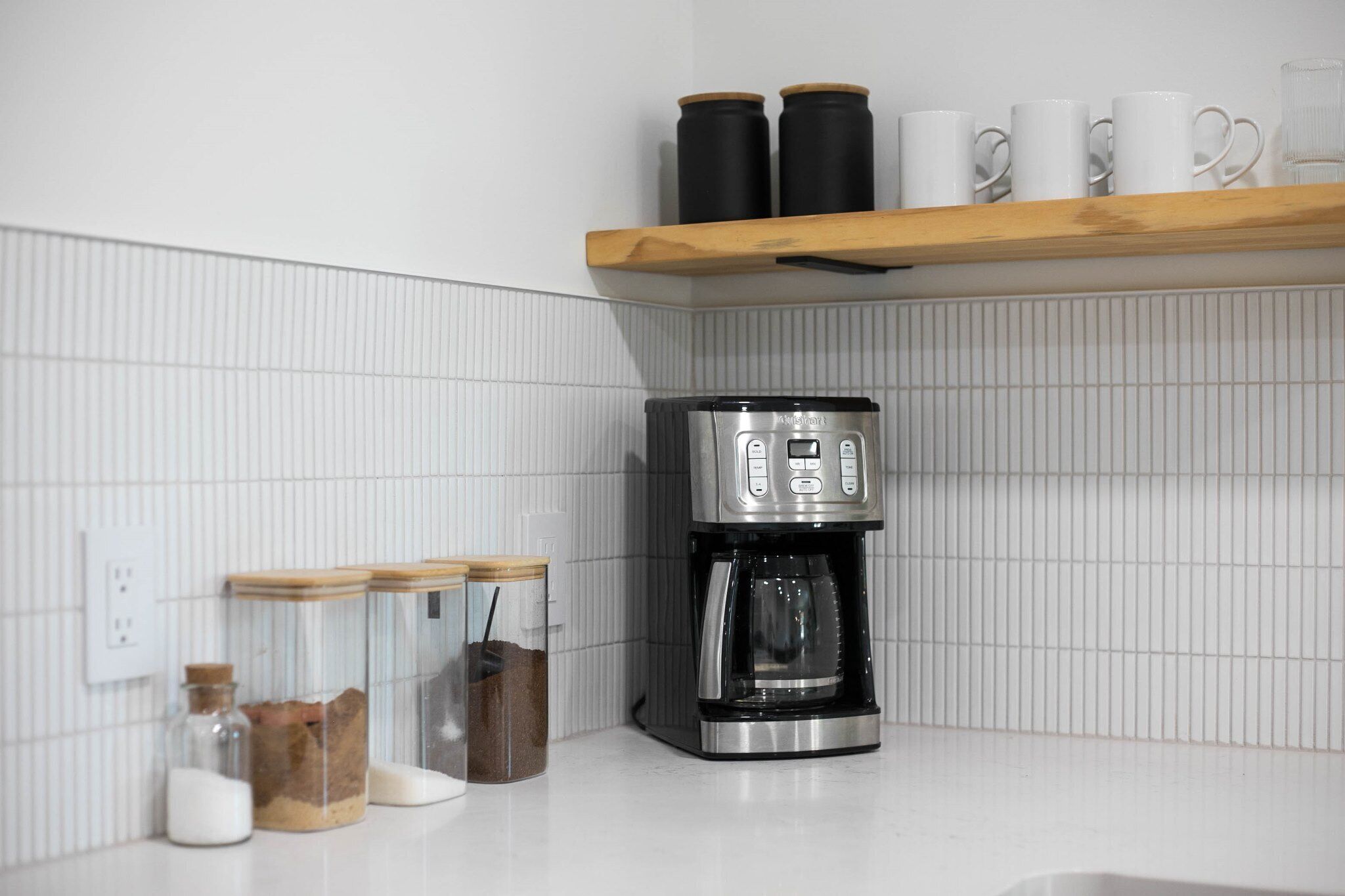 Une cuisine équipée d’une machine à café, de bocaux et d’une étagère où sont rangées des tasses.