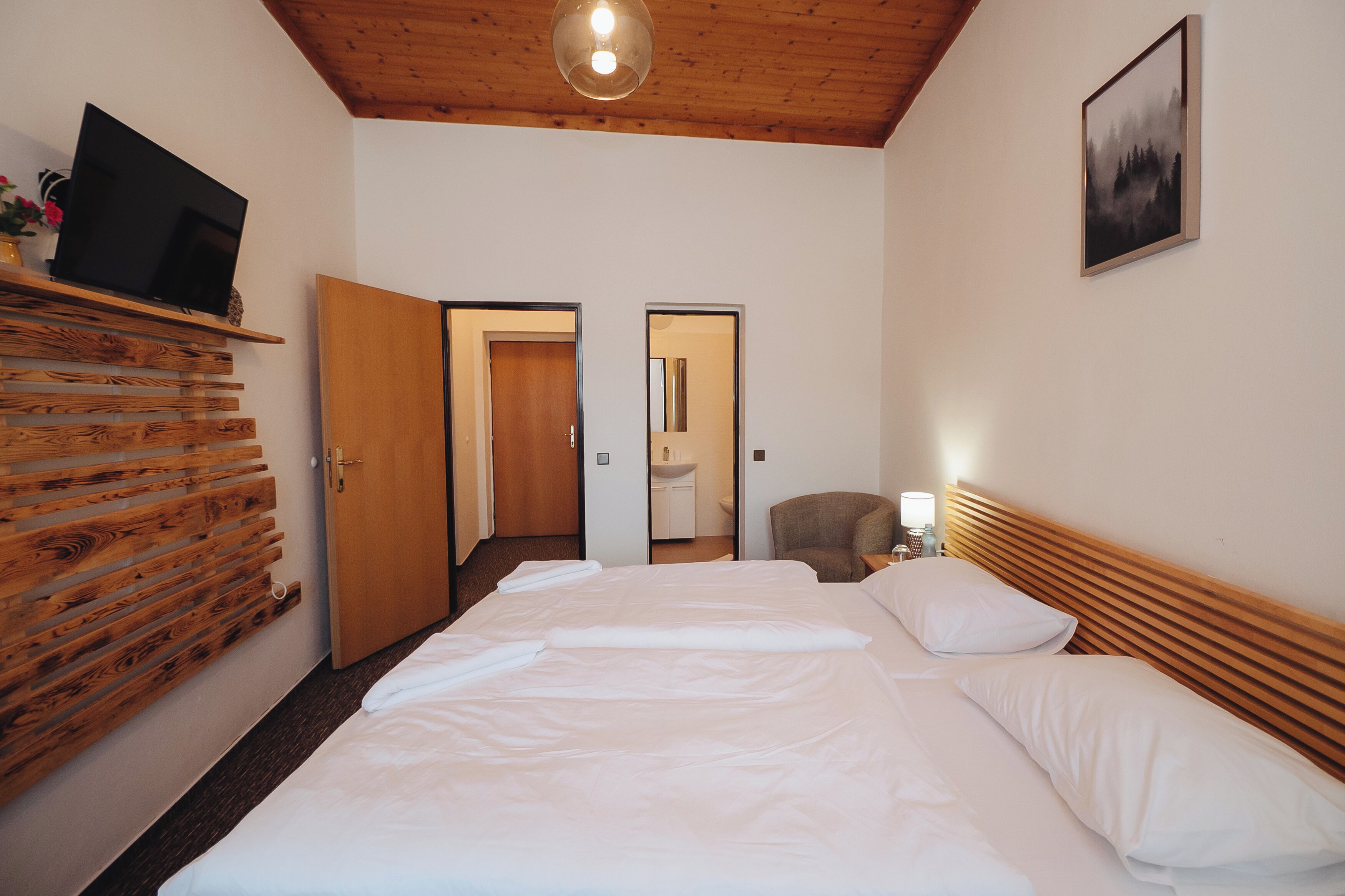 Habitación de hotel con una cama grande, cabecero de madera, un televisor de pantalla plana sobre una estantería, un cuadro en la pared y una puerta que da al baño.